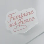 Wavy vinyl sticker with cursive pink text reading “Feminine and Fierce” above a small illustrated cloud. Features partial peel to showcase adhesive strength. Printed on clear vinyl with soft contour edging. Designed by PuffSticker.com as part of an empowerment-themed sticker collection for journals, laptops, and branding kits.