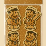 Packaged sheet of puffy stickers labeled “PUFFY STICKERS” featuring four cartoon stickers of Fred Flintstone from The Flintstones. Each sticker shows Fred in a different pose: hands on hips, waving, smiling, and arms crossed. Set against a brown polka dot background with white header text. Perfect for retro-themed crafts, cartoon memorabilia, and playful sticker collections.