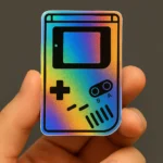 Holographic vinyl sticker featuring a retro handheld gaming console design inspired by the classic Game Boy. The sticker showcases a rainbow iridescent background with black outlines of the screen, directional pad, A and B buttons, and speaker grille. Held between two fingers against a dark background, the sticker reflects vibrant colors depending on the light angle. Ideal for nostalgic gamers, tech-themed sticker sets, and retro-inspired stationery. Designed by PuffSticker.com