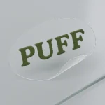 Clear vinyl label with bold green uppercase text reading “PUFF” partially peeled from a smooth surface to demonstrate adhesive quality. Designed for minimalist packaging, custom branding, and waterproof durability. Printed by PuffSticker.com with precision to maintain clarity and crisp edges. Ideal for cosmetic bottles, product pouches, or stationery branding.